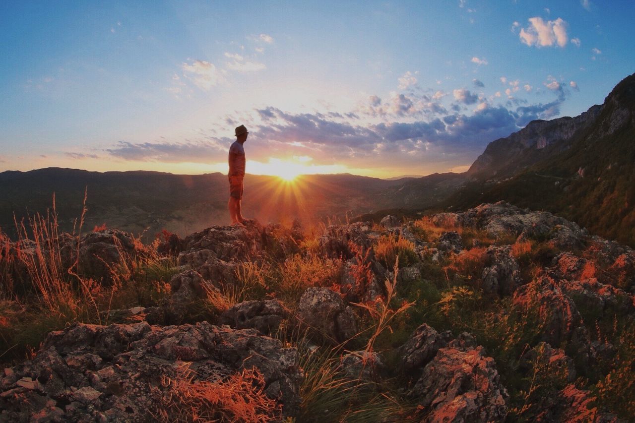 Side view of man standing on cliff against mountains during sunset
