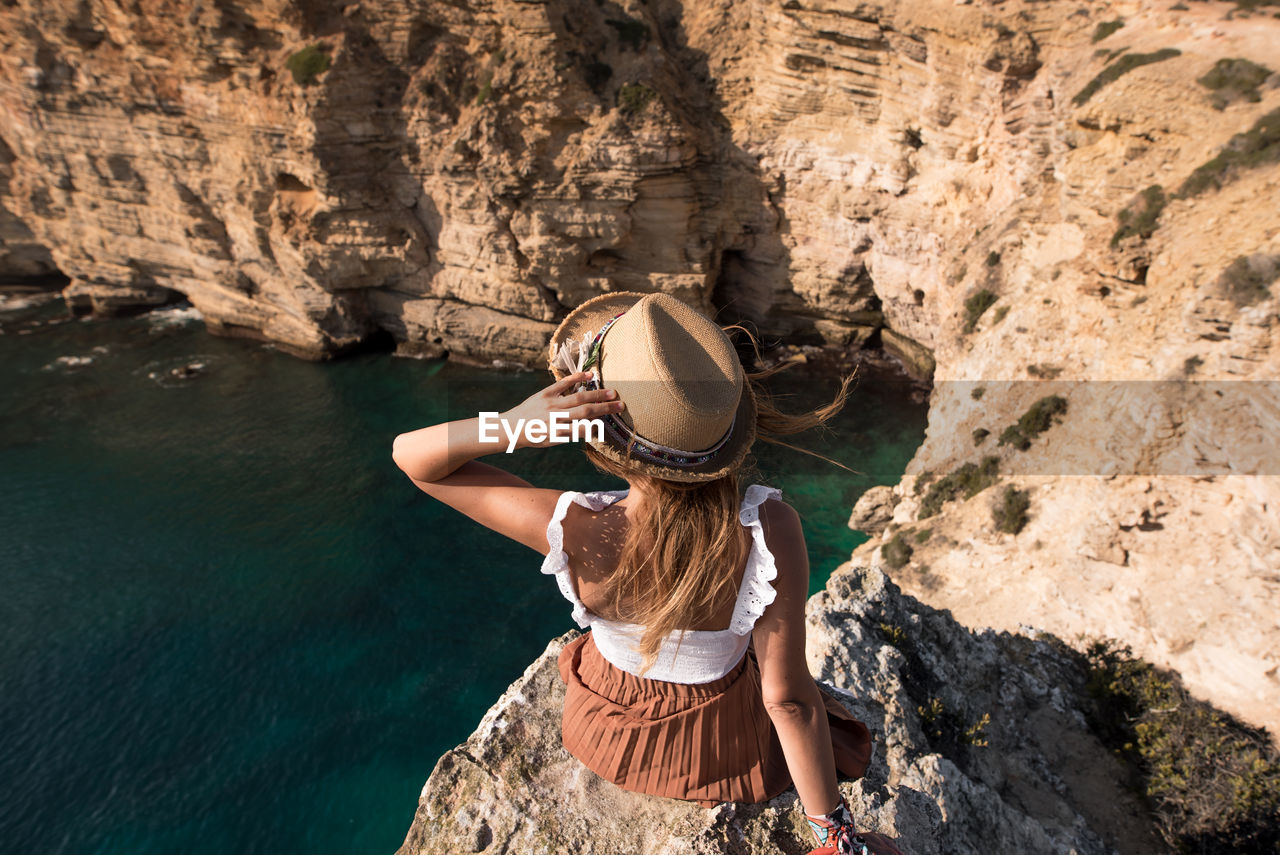 Back view of unrecognizable young woman in summer clothes and straw hat sitting in cliff admiring landscape at cape saint vincent in sagres, portugal