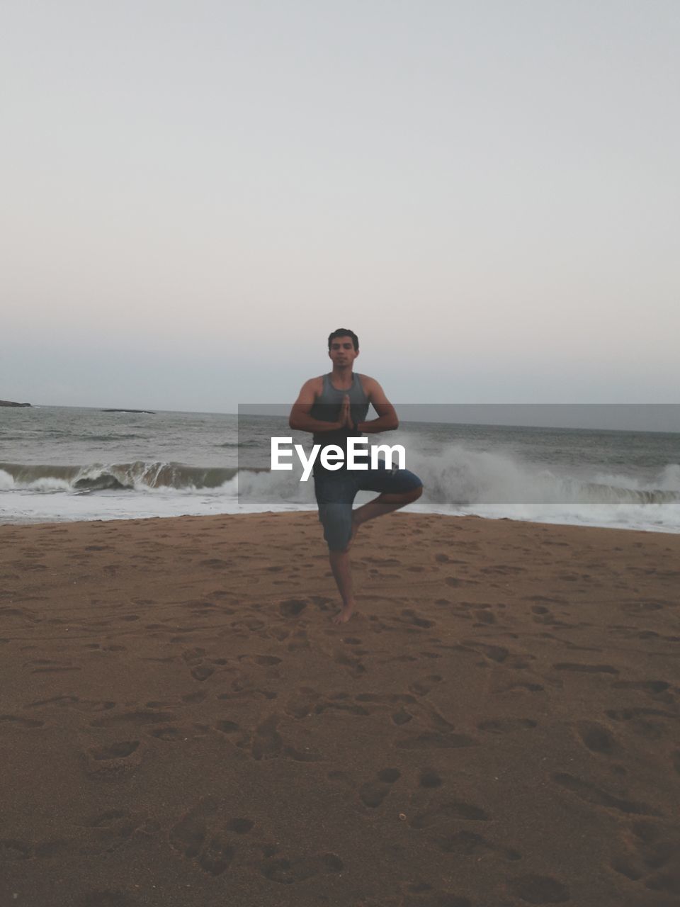 Full length of man doing tree pose yoga while exercising on shore at beach against sky