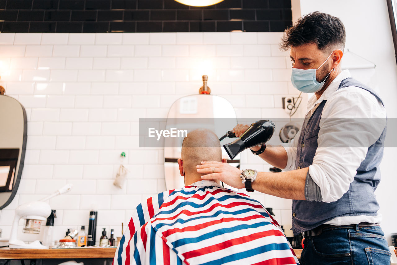 Anonymous male stylist in mask using hairdryer while touching bald head of man in striped cloth in beauty salon