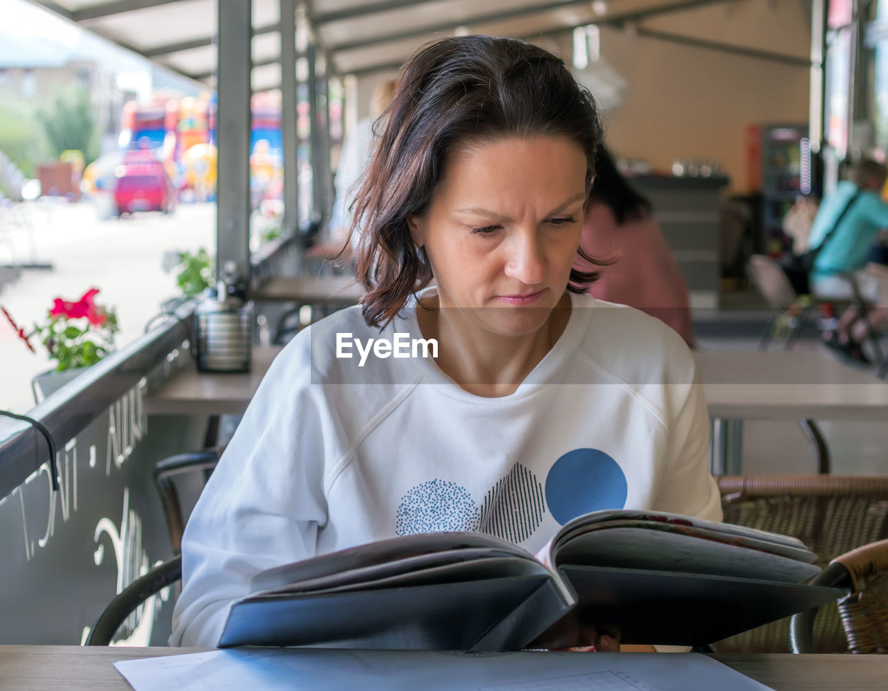 Caucasian middle-aged woman is choosing a dish at a menu in a street cafe.