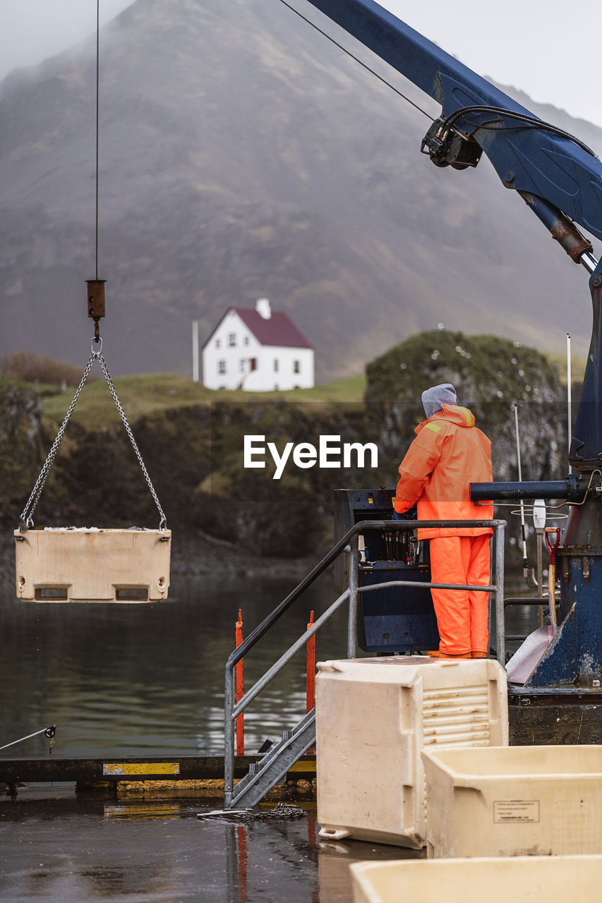 Anonymous man in orange uniform controlling crane during work on fish farm on gray day in mountains of iceland