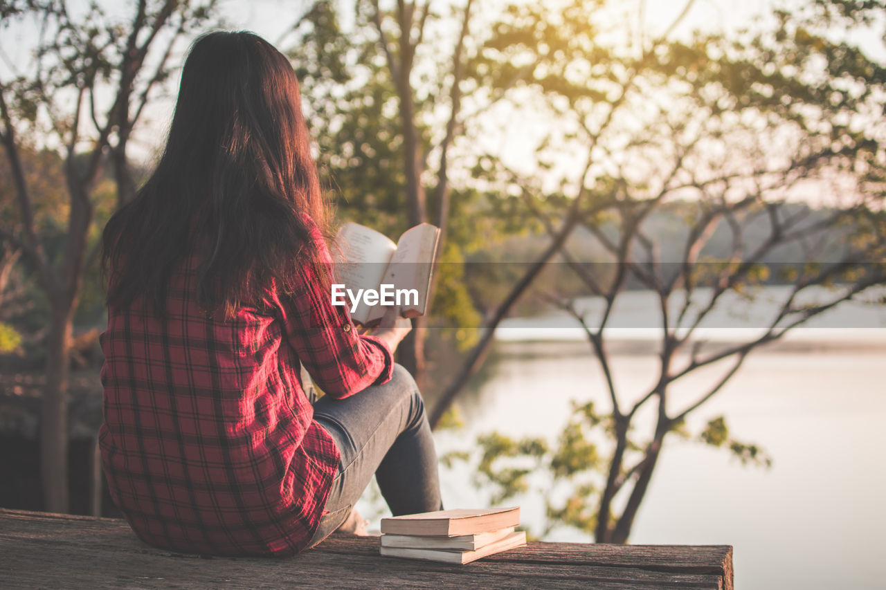 Rear view of woman reading book while sitting by lake