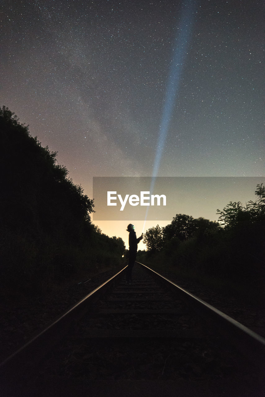 Silhouette person with illuminated flashlight standing on railroad track against sky at night