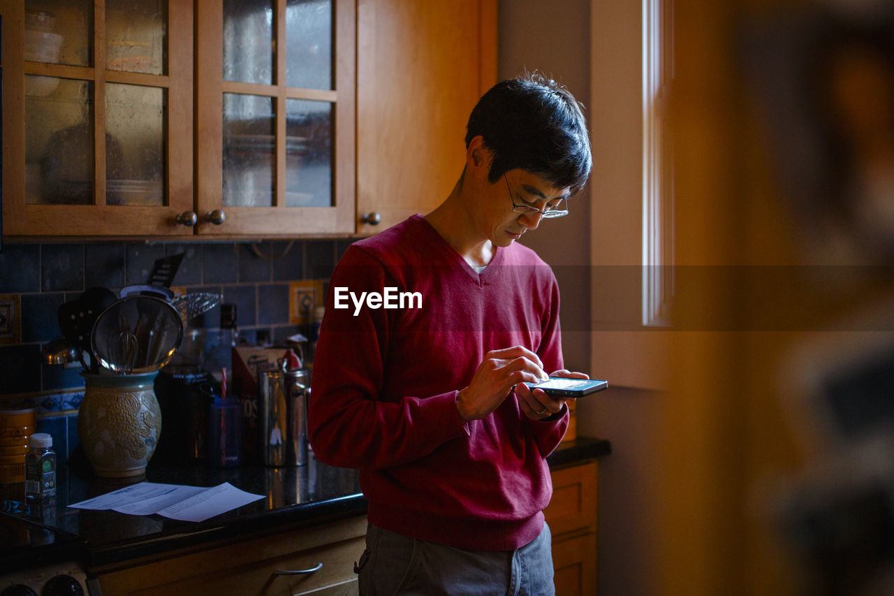 A man stands in a kitchen by window light texting on a cell phone