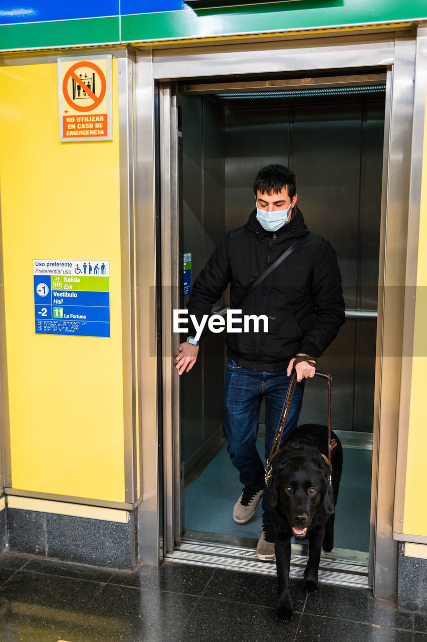Full body of male with blindness in medical mask calling elevator standing with guide dog