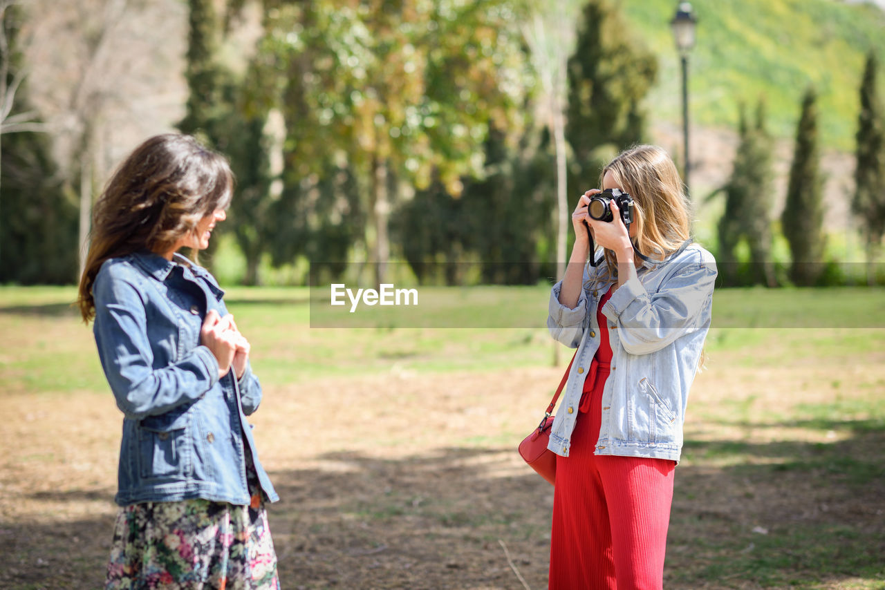 Woman photographing smiling friend in park during sunny day