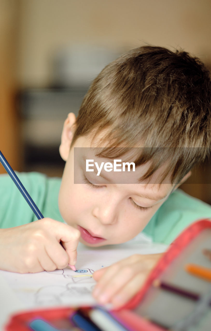 Cute and smiling cucasian boy draws with colored pencils while sitting at the table