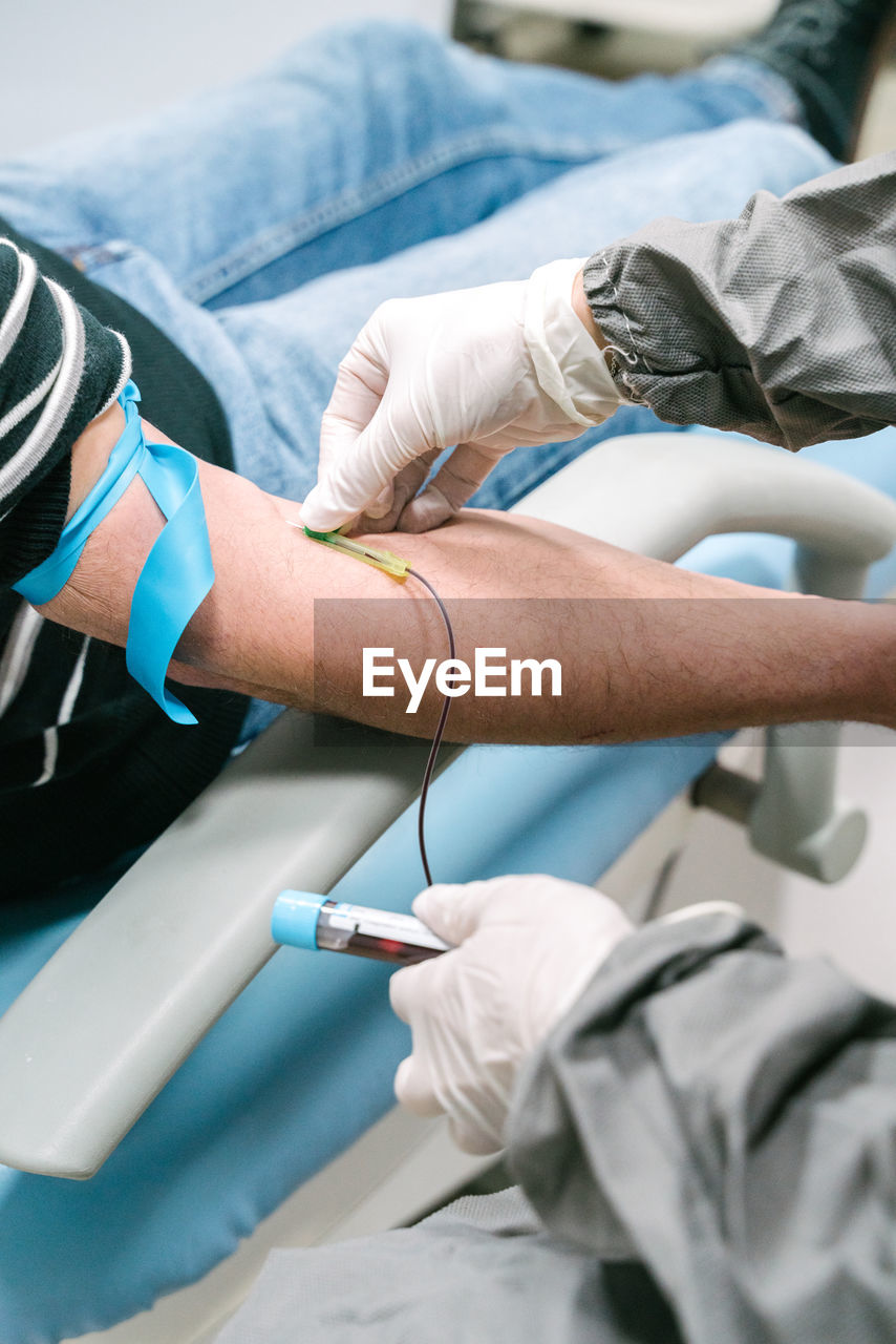 Professional female medical specialist in protective uniform preparing arm of anonymous patient for blood test in modern lab