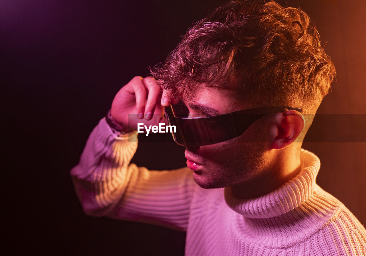 Side view of self assured young male in stylish outfit adjusting futuristic sunglasses while standing in dark studio with neon illumination