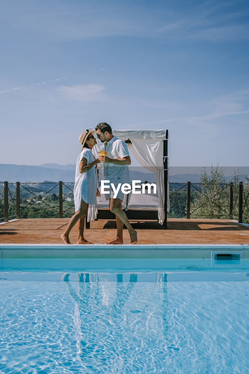 Couple standing by swimming pool against sky