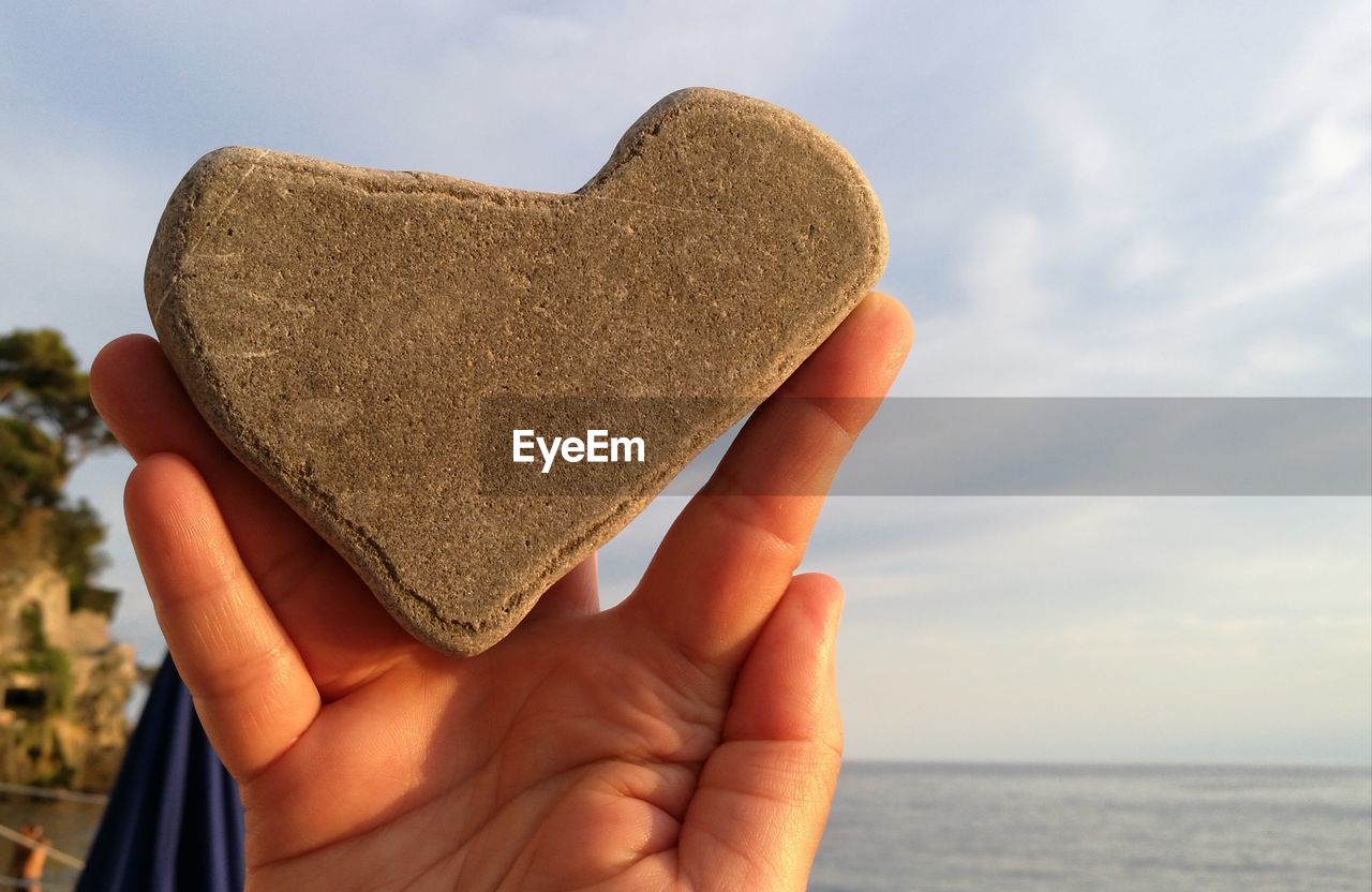 Close-up of hand holding heart shape rock against sky