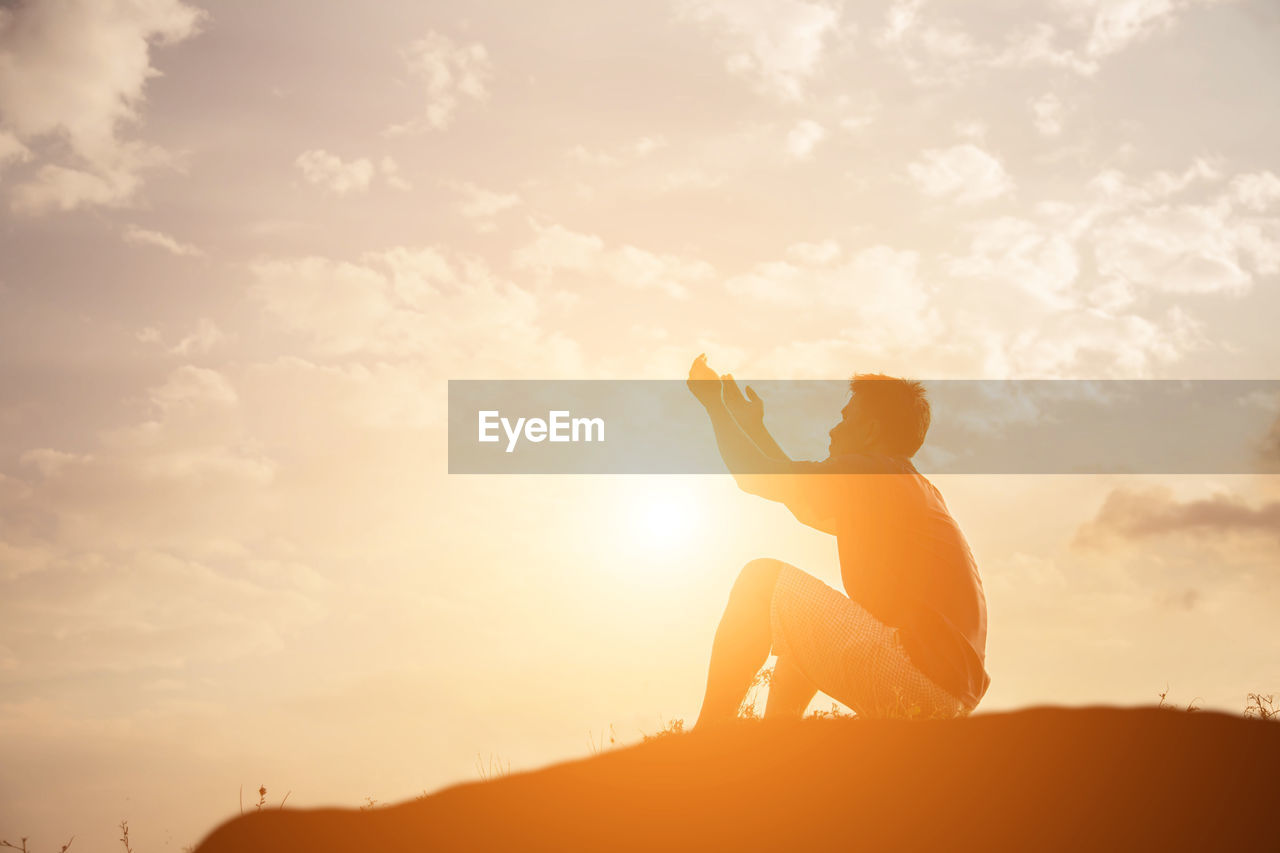 Side view of man gesturing while sitting on land against sky during sunset