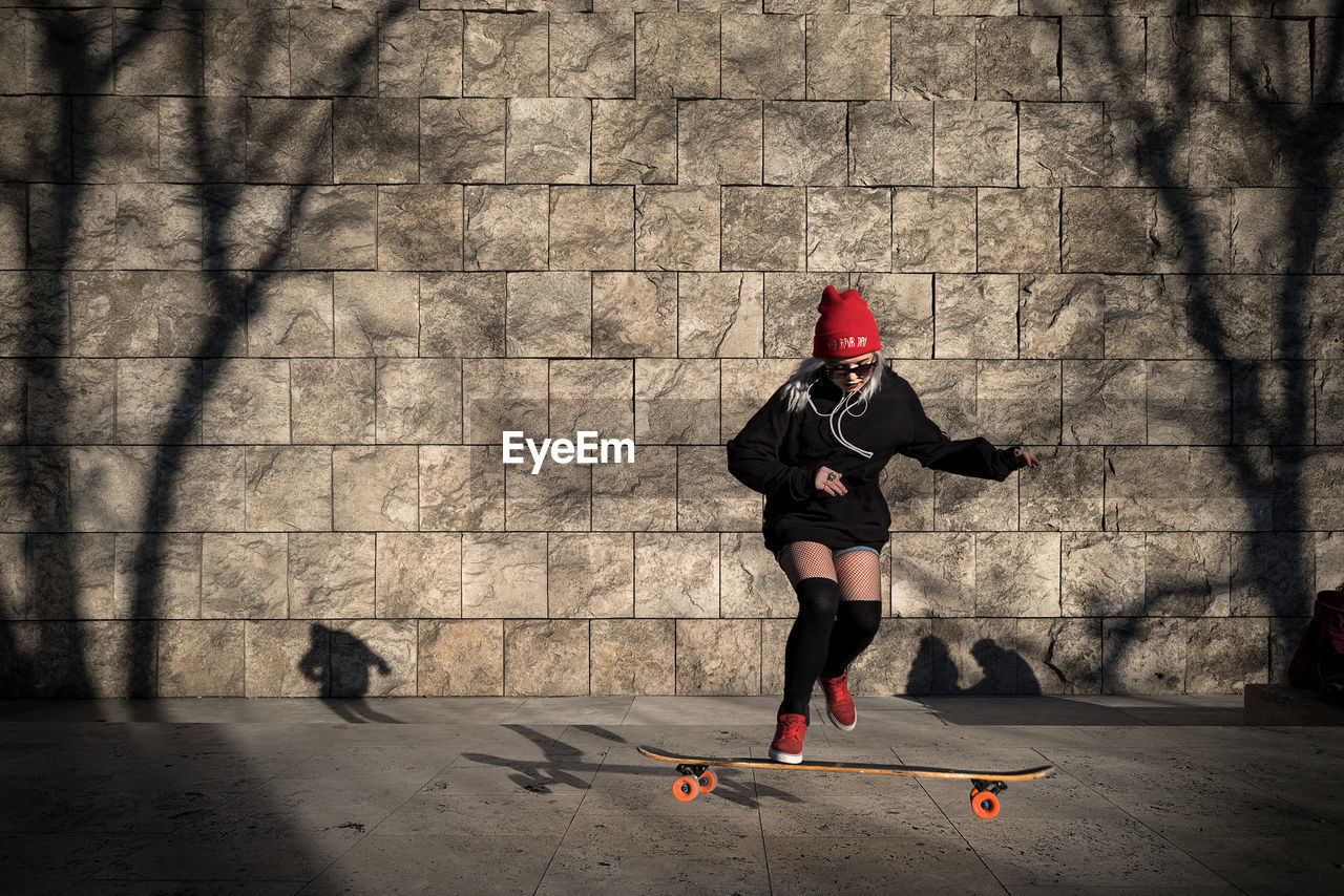 Full length of woman skateboarding on footpath against wall