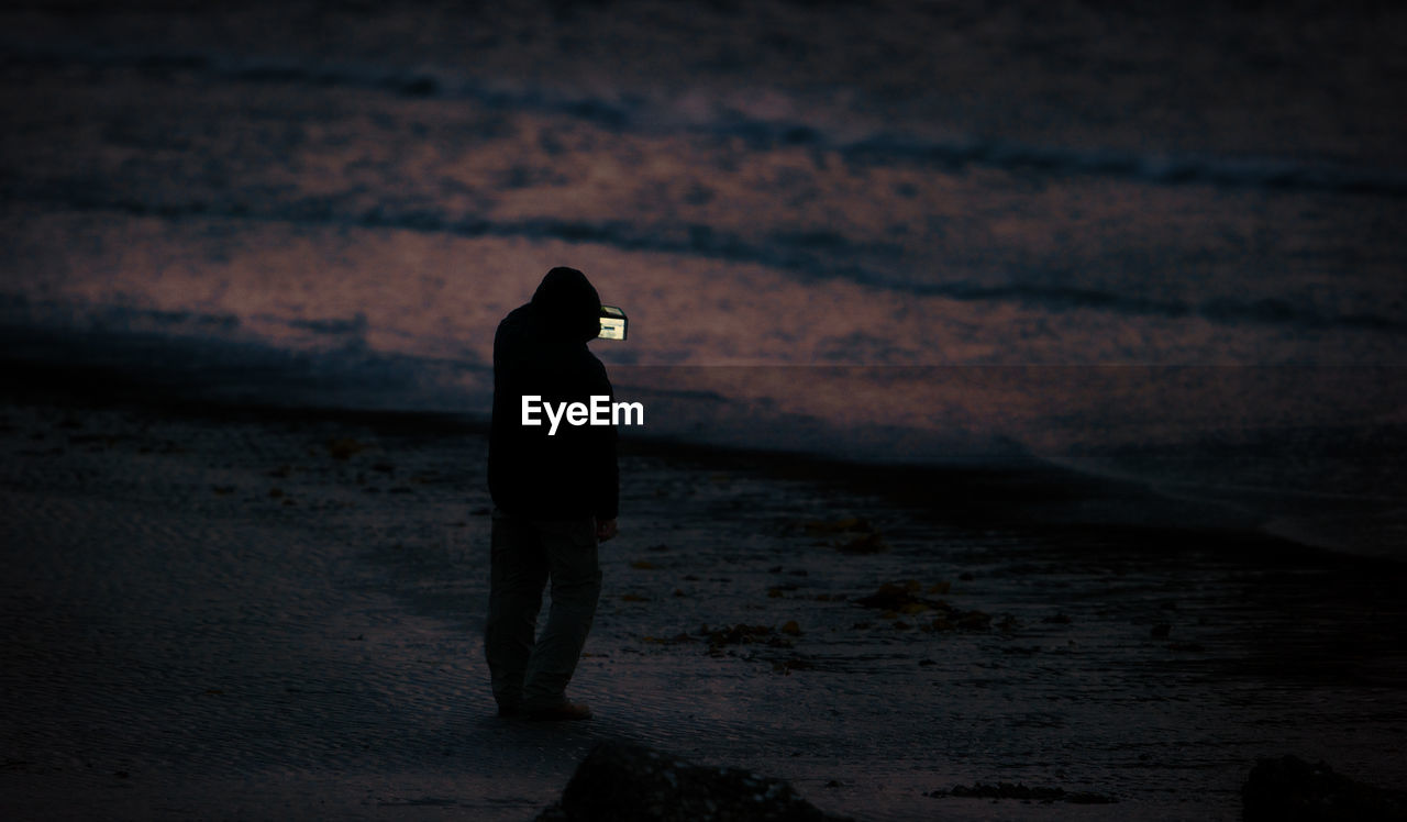 Silhouette person photographing with camera while standing on beach at sunset