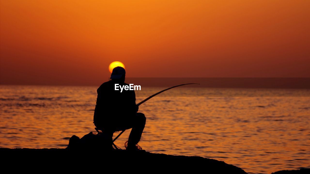 Silhouette man fishing in calm sea at sunset