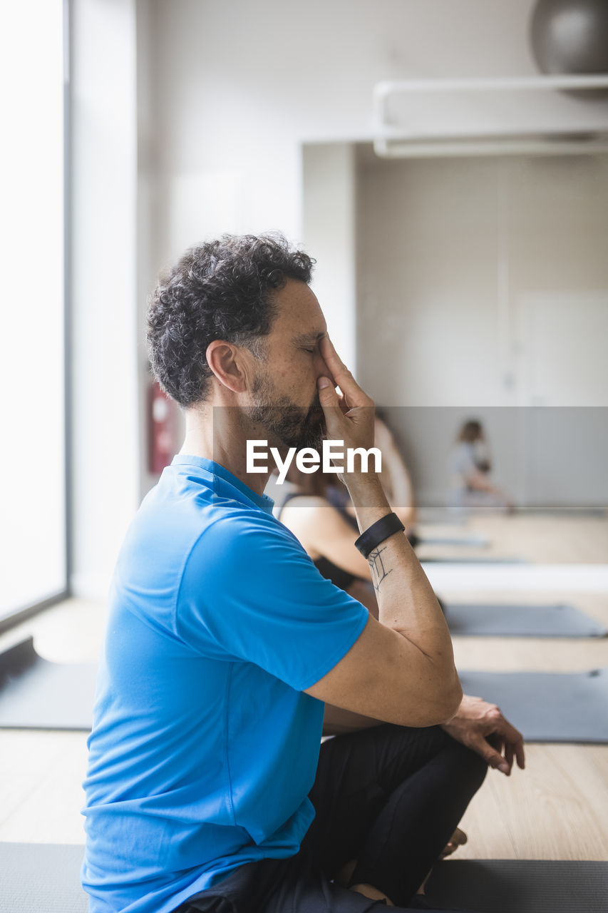 Concentrated man in activewear sitting with eyes closed and legs crossed and keeping fingers in nasarga mudra gesture while practicing breathing exercise during yoga lesson in studio