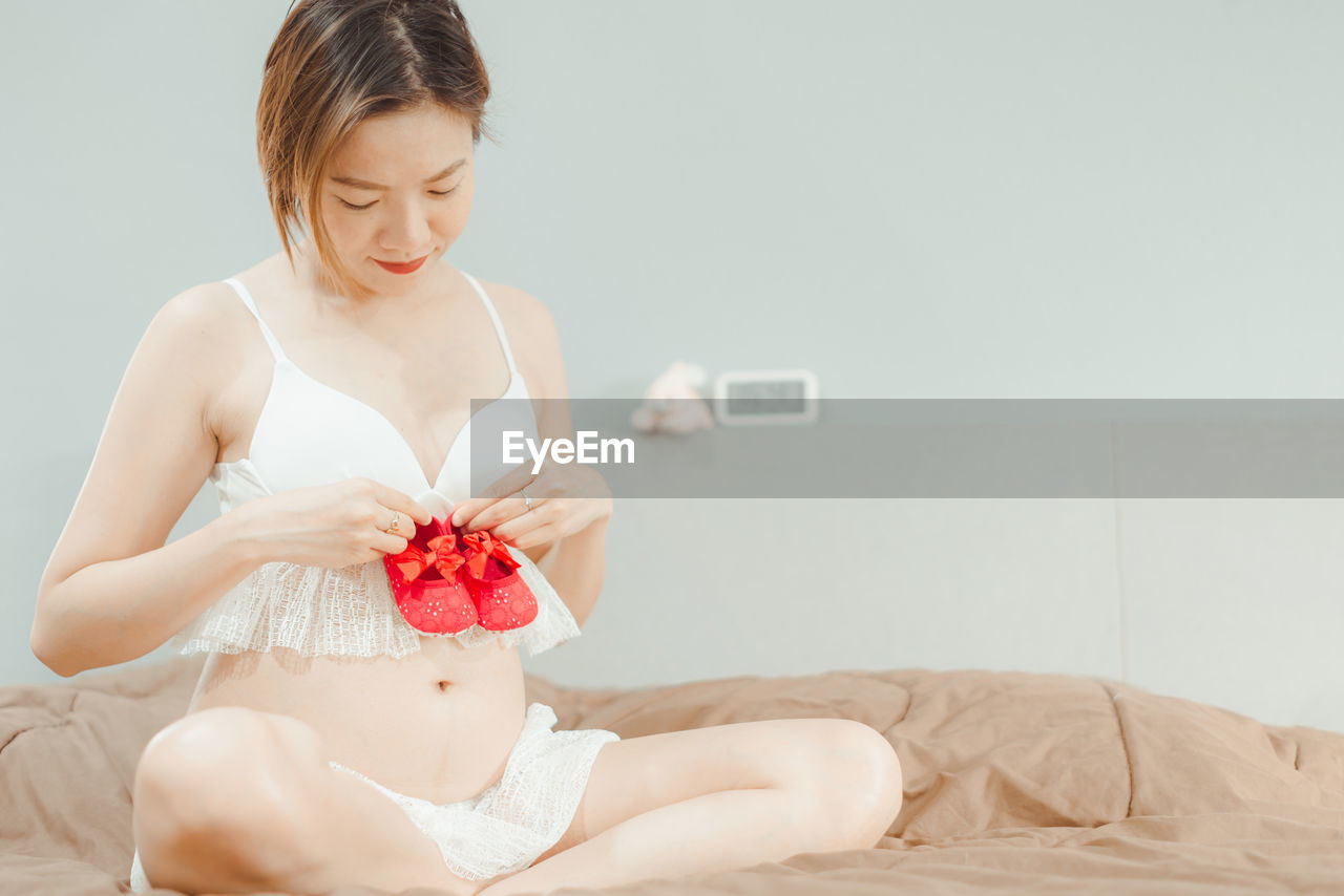 Pregnant woman holding baby booties against abdomen while sitting on bed at home