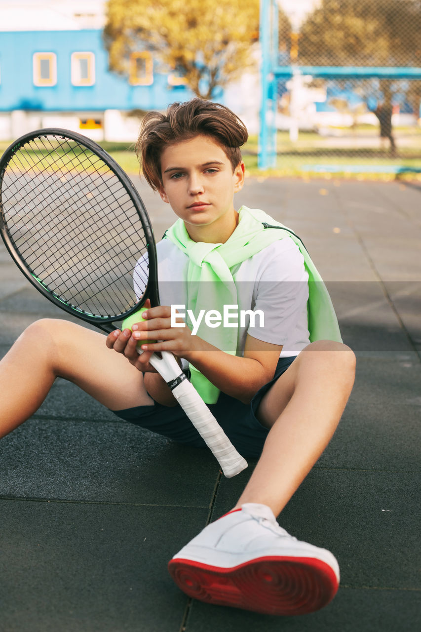 Portrait of boy with tennis racket sitting in court