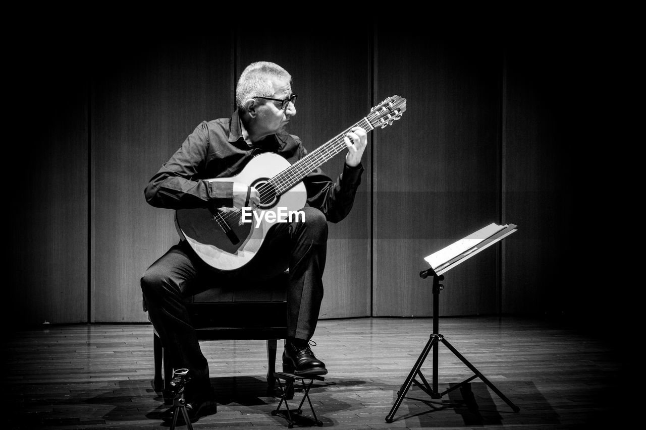 music, musical instrument, arts culture and entertainment, black and white, musician, black, adult, monochrome, performance, men, one person, indoors, monochrome photography, entertainment, musical equipment, string instrument, full length, performance art, stage, sitting, guitar, skill, white, person, seat, performing arts event, concert, performing arts, copy space, plucking an instrument, plucked string instruments, chair