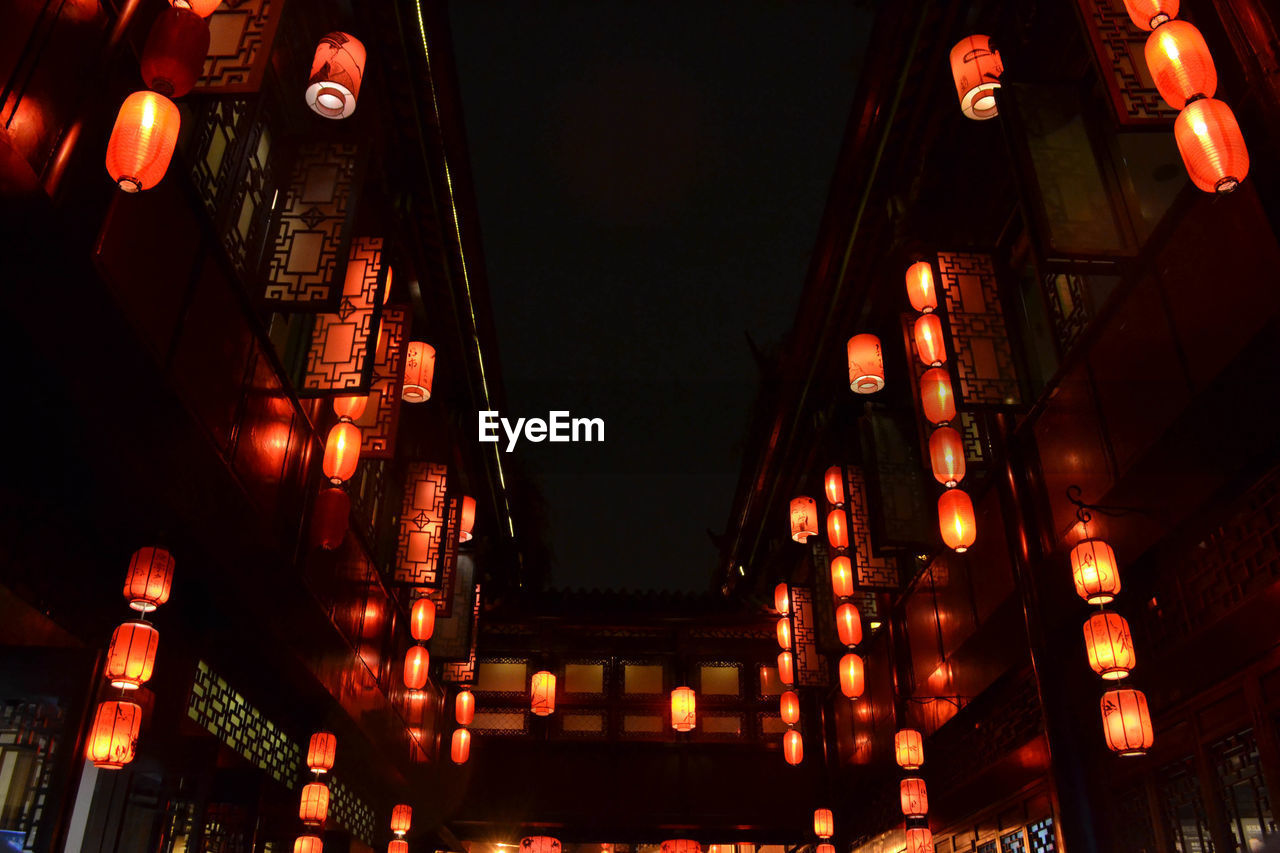 LOW ANGLE VIEW OF ILLUMINATED LANTERNS HANGING AMIDST BUILDINGS IN CITY