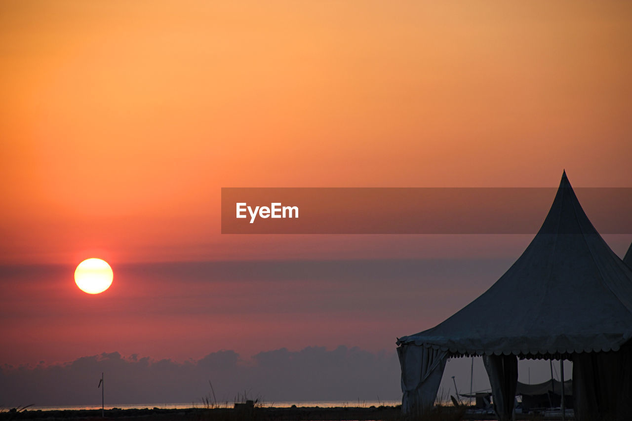 Silhouette tent at beach against orange sun in romantic sky