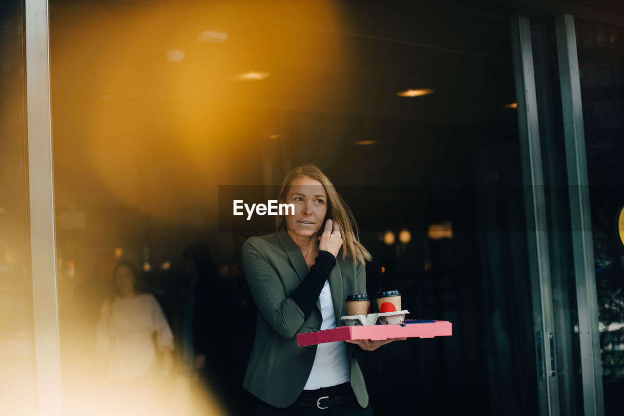 Mature businesswoman talking on smart phone while holding food and drink against cafe