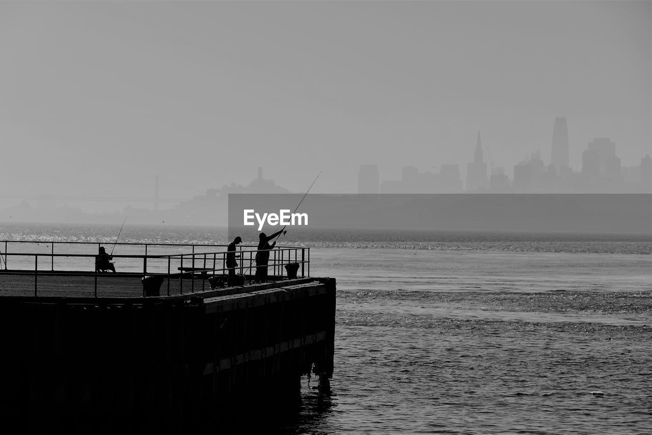 Silhouette people fishing on pier at sea against sky in city