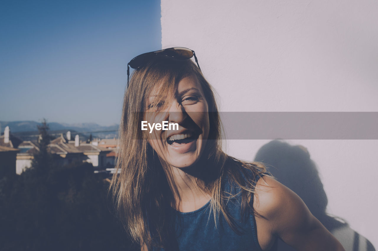 Portrait of happy woman laughing by wall against clear sky