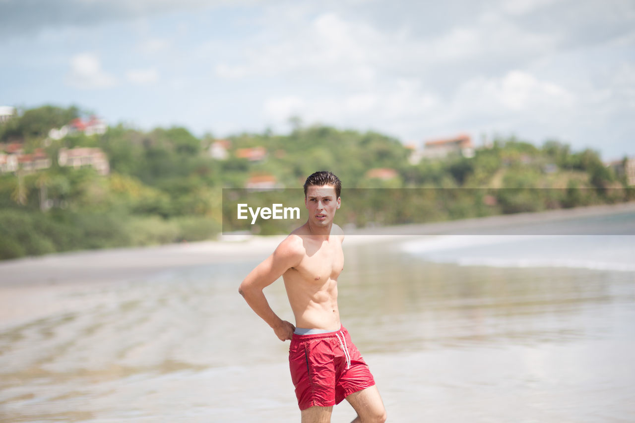 Shirtless young man looking away while standing at shore