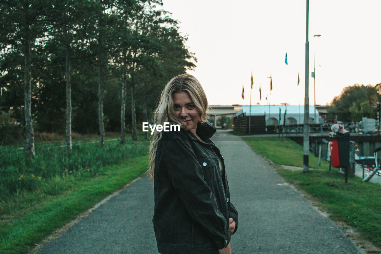 PORTRAIT OF SMILING YOUNG WOMAN STANDING BY ROAD