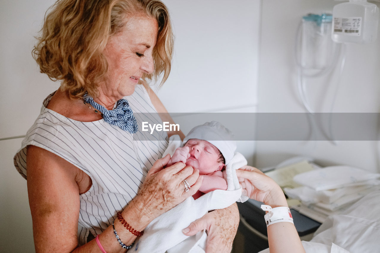 Happy mature female greeting newborn grandchild in hospital ward while young mother lying in bed and receiving medical treatment