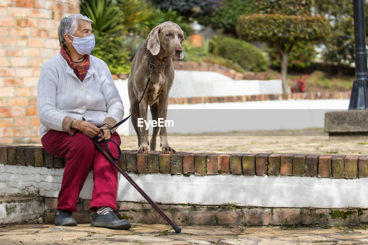 Full length of senior wearing mask with dog sitting outdoors