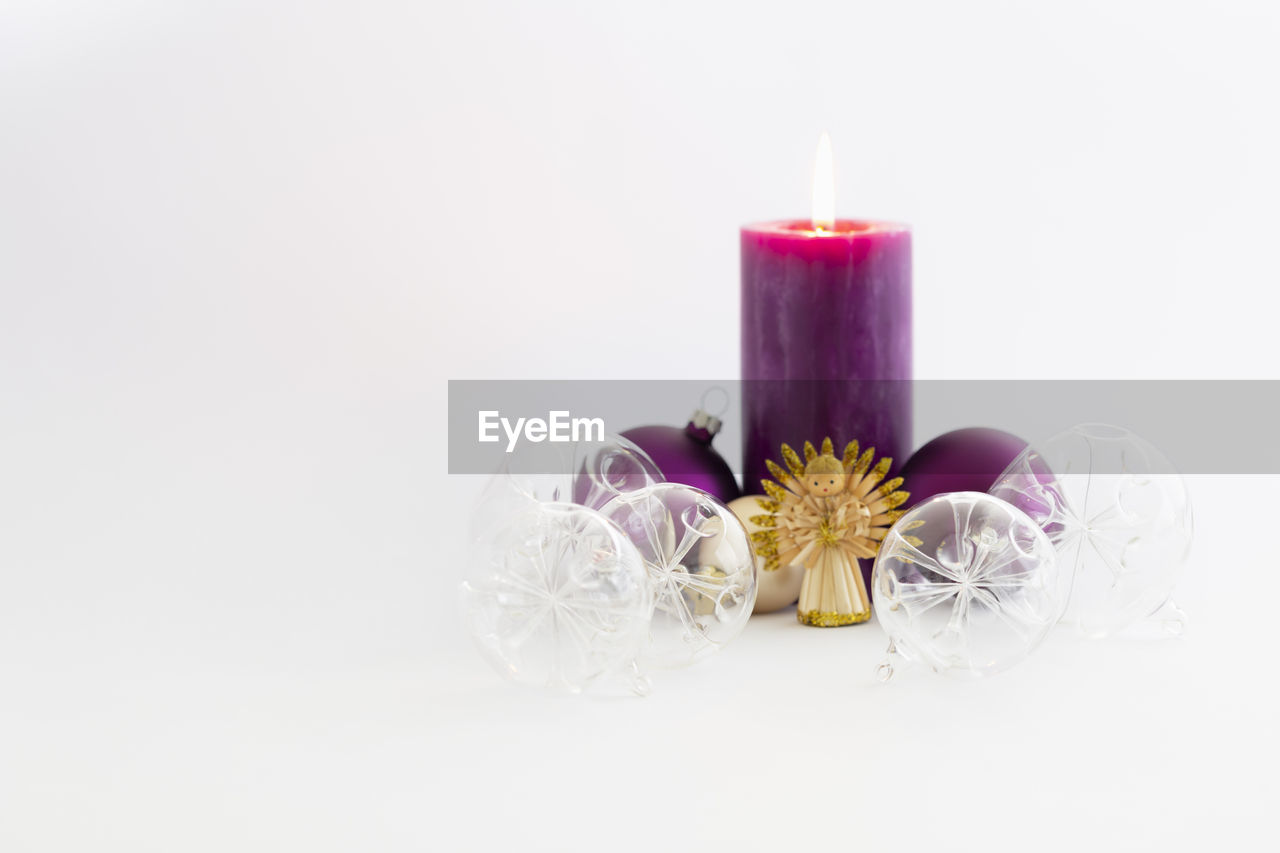 Transparent christmas glass balls, a purple  burning candle and a straw angel on white background