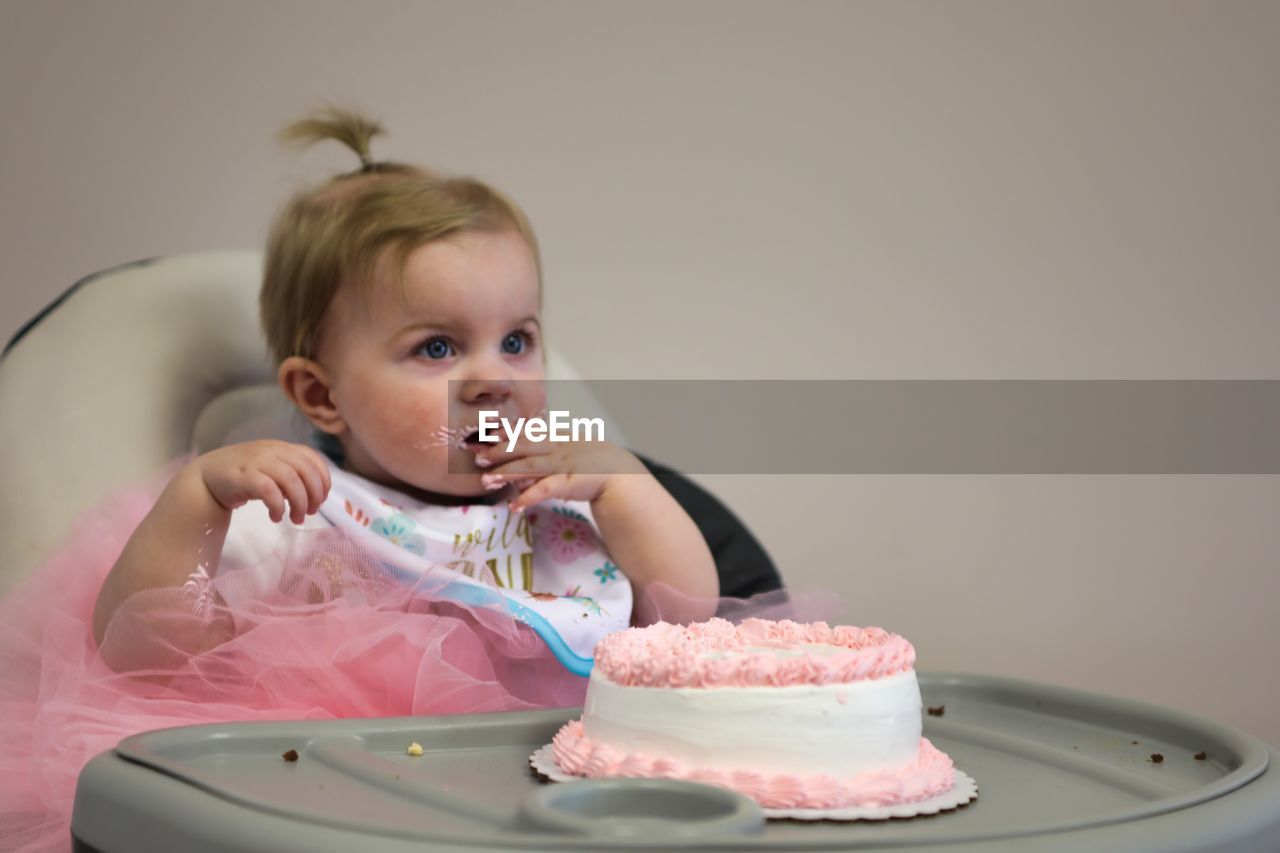 Cute baby girl eating birthday cake while sitting on high chair at home