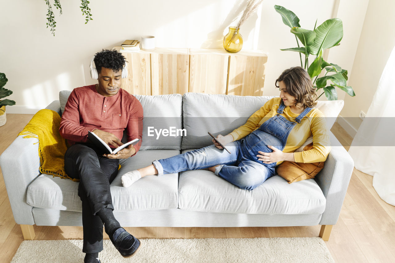 Husband listening to music and reading a book with his pregnant wife who is watching a tablet on a sofa. interracial couple concept