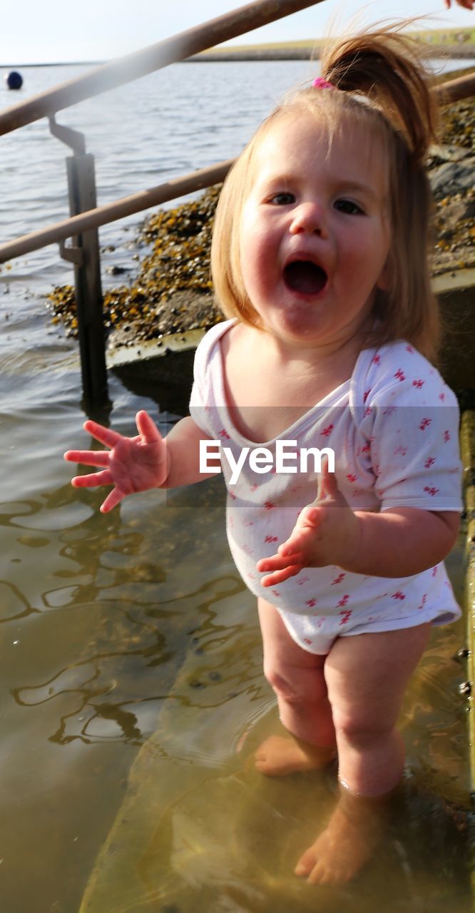 child, childhood, water, one person, emotion, baby, innocence, cute, nature, portrait, mouth open, fun, day, blond hair, happiness, female, women, front view, smiling, full length, clothing, toddler, person, looking at camera, outdoors, vacation, enjoyment, cheerful, sea, leisure activity, human mouth, lifestyles, looking, standing, holding, positive emotion, facial expression, carefree, holiday, laughing