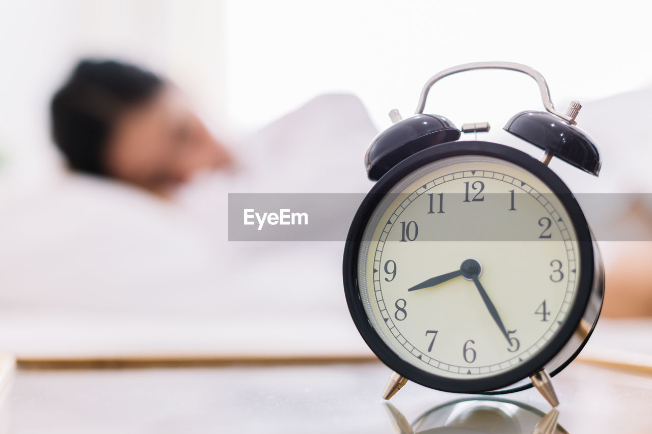 Close-up of alarm clock on side table with woman sleeping in background