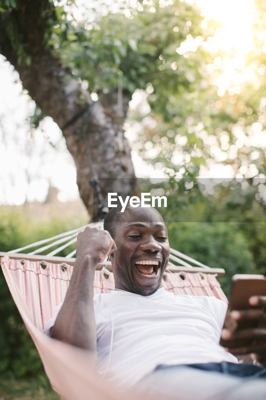 Cheerful mid adult man looking at mobile phone while relaxing on hammock in backyard