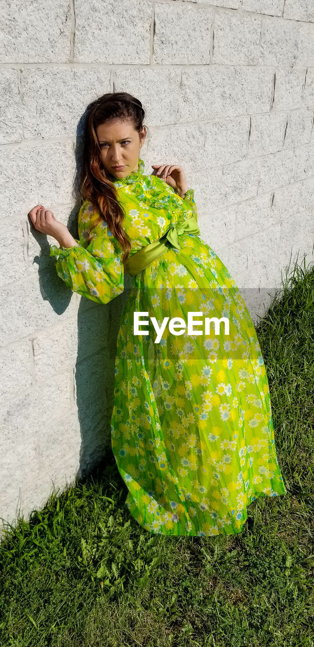 Full length of woman in green floral dress leaning on stone wall