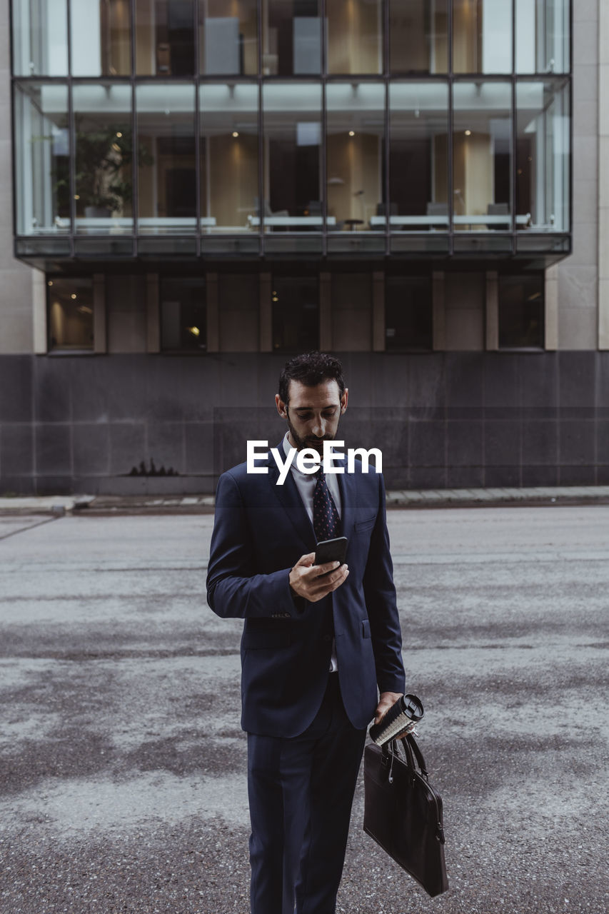 Male entrepreneur with bag using smart phone while standing in city