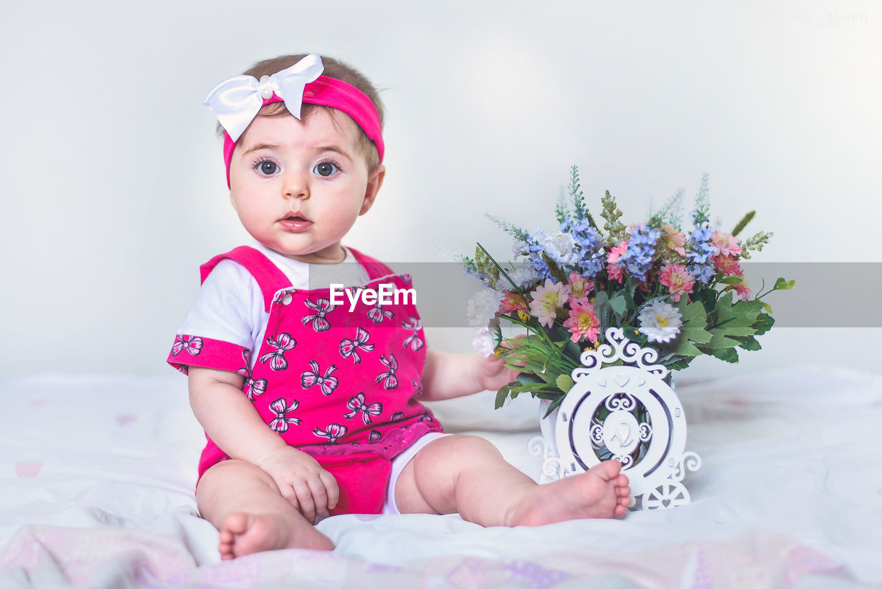 Portrait of cute baby girl with flowers sitting on bed at home