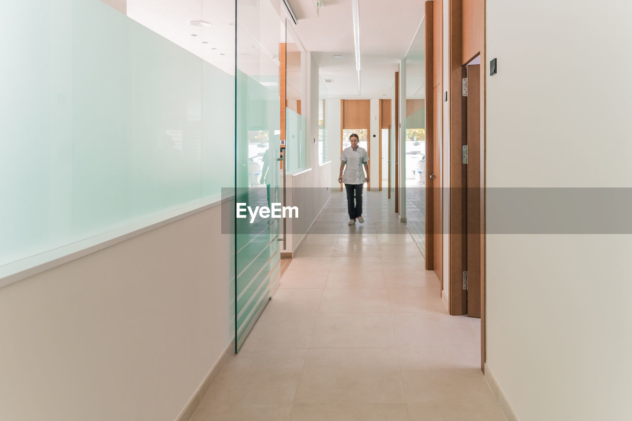 Full body woman in medical uniform looking away while standing in hallway of modern clinic during work