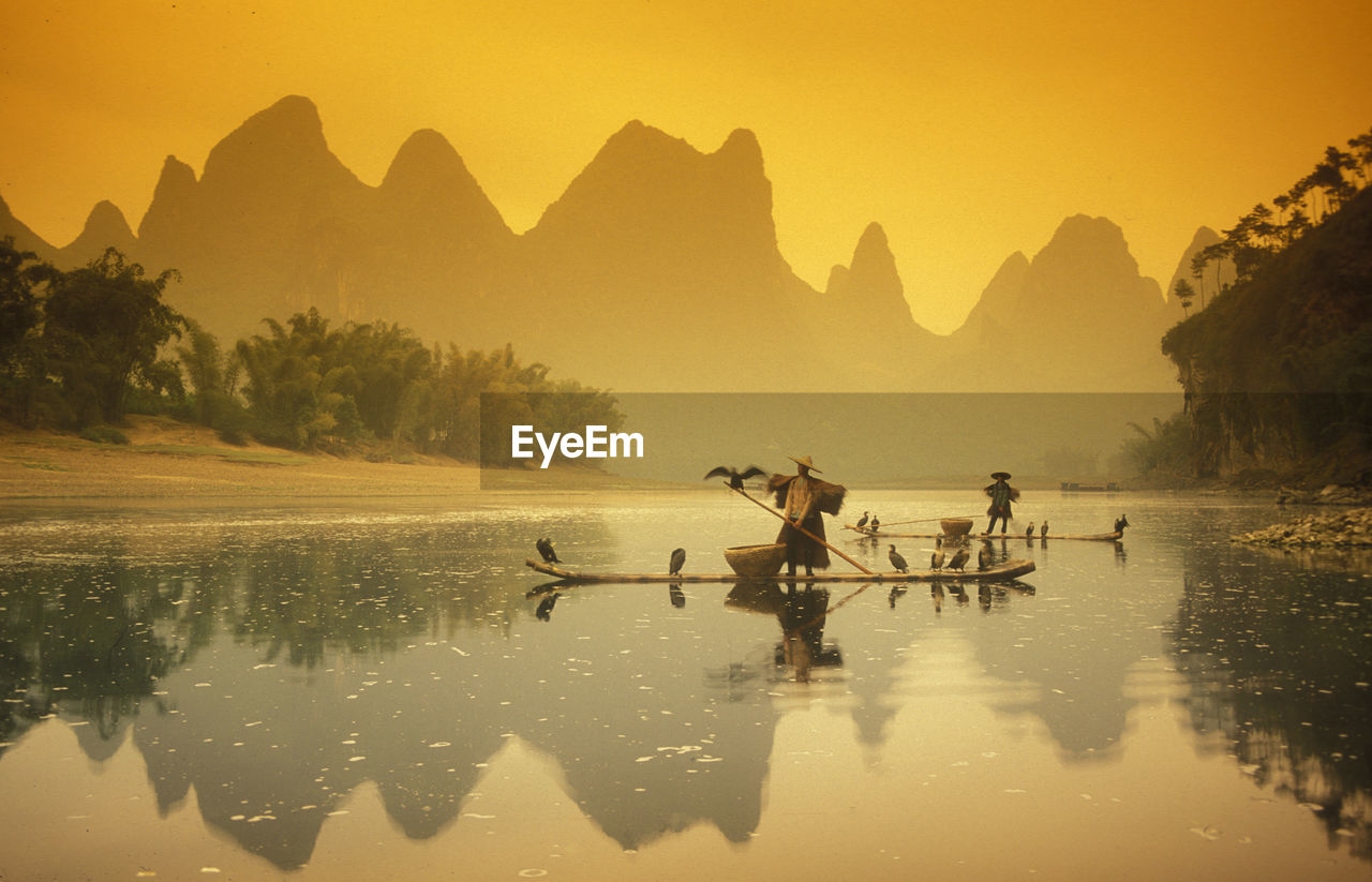 People with birds on boat in lake against mountains during sunset