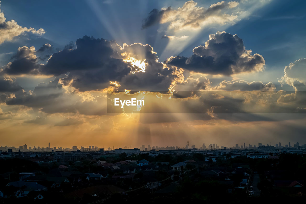 LOW ANGLE VIEW OF SUNLIGHT STREAMING THROUGH CLOUDS OVER CITYSCAPE