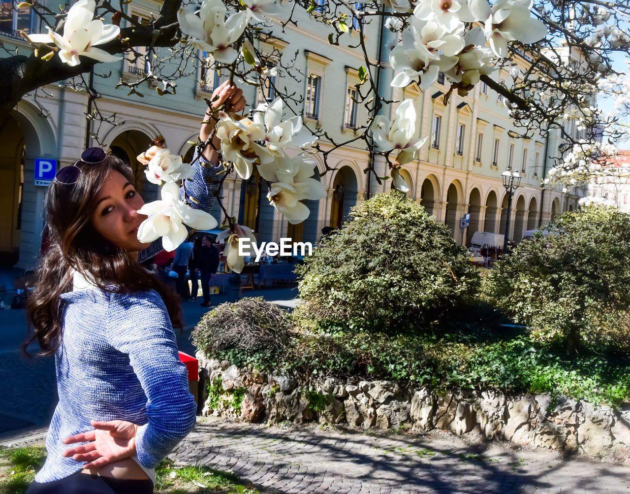 WOMAN STANDING BY FLOWERING PLANTS AGAINST BUILDING