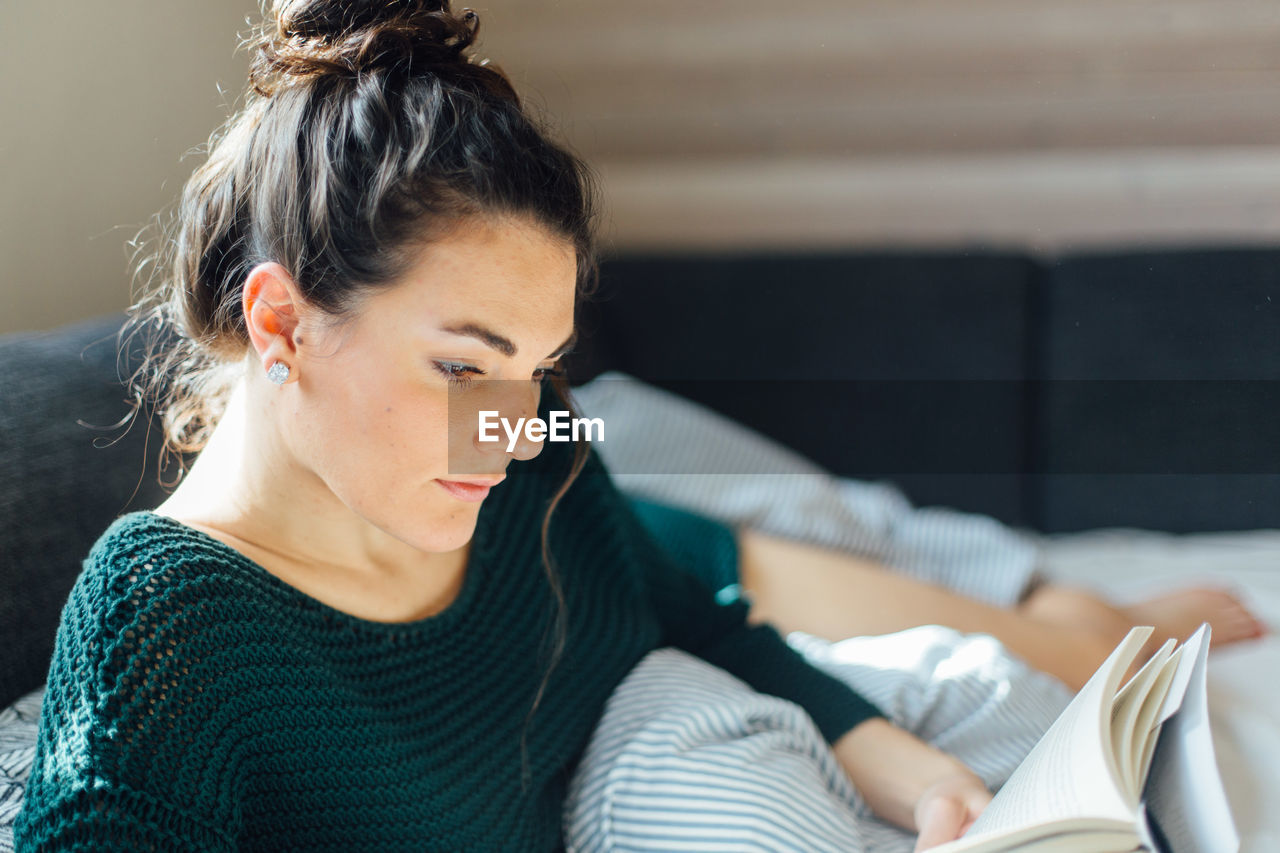 Young woman reading book while lying on bed at home