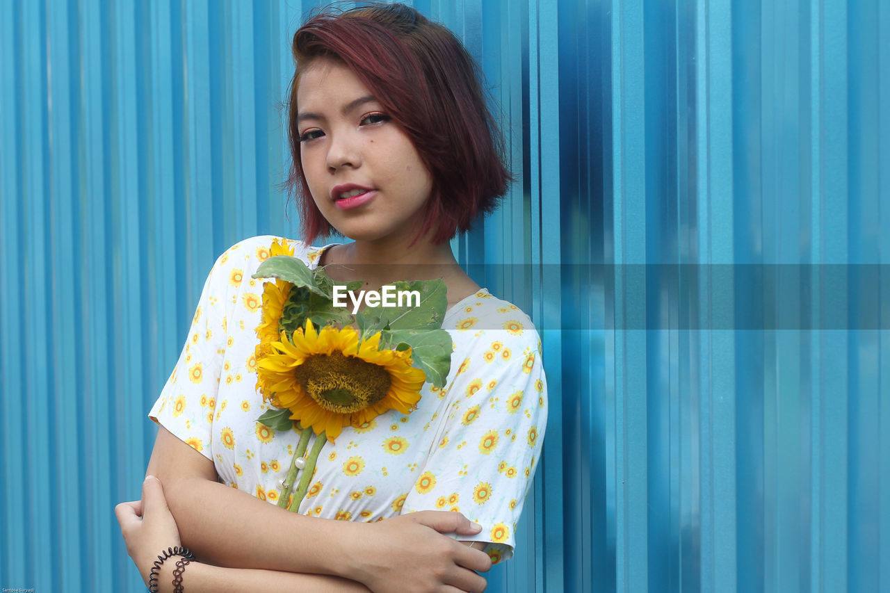 Portrait of woman holding sunflowers while leaning on corrugated iron