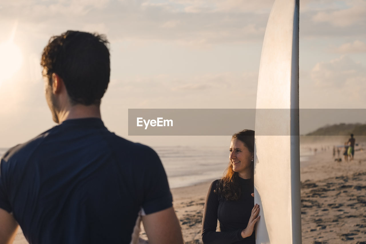 Young man with female friend holding surfboard at beach during sunset