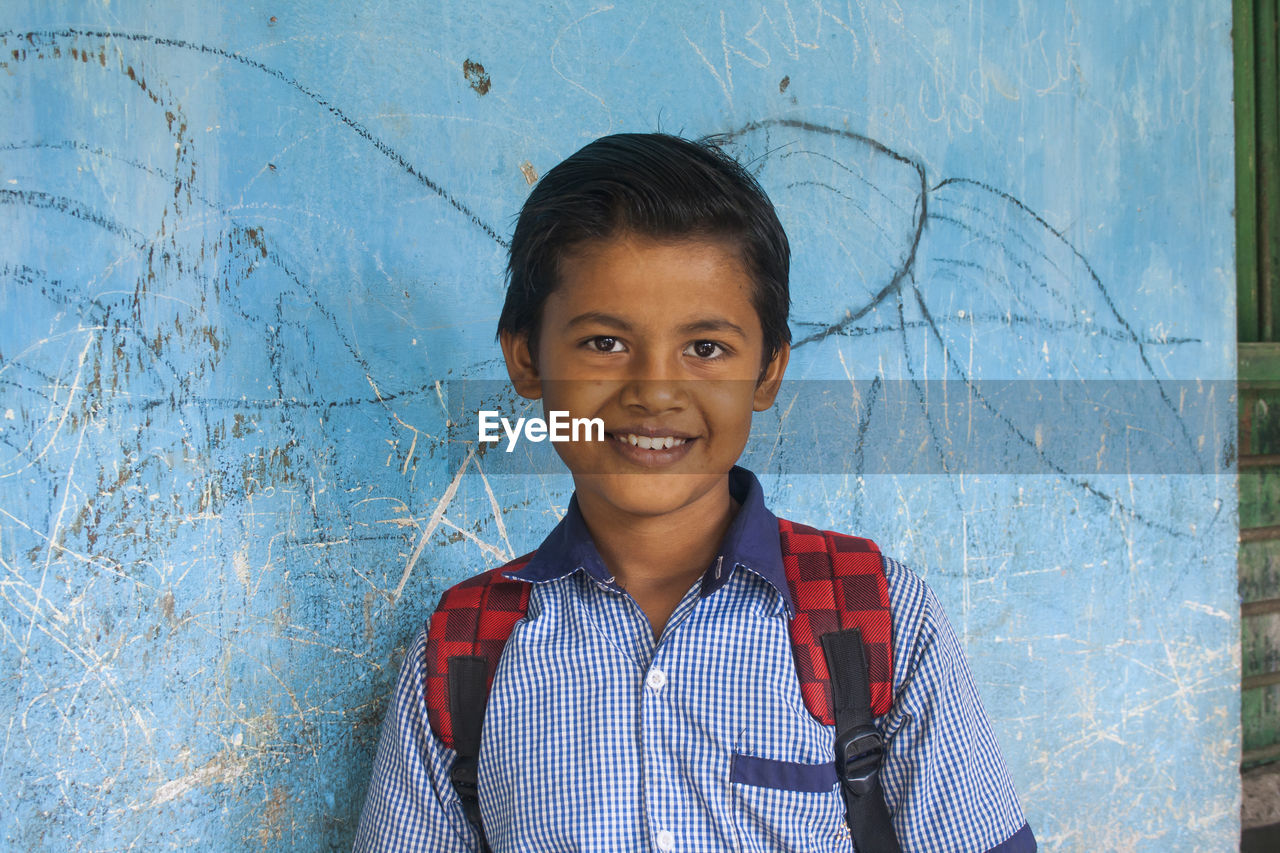 PORTRAIT OF SMILING BOY STANDING AGAINST BLUE WALL